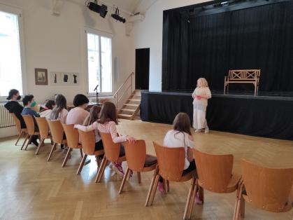Mädchen* sitzen auf Stühlen vor einer Bühne und hören der Theaterpädagogin zu.